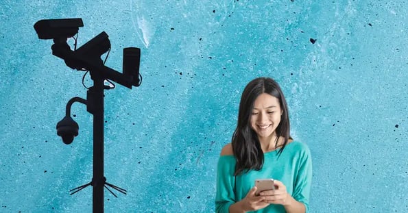 A surveillance camera looms behind a young Asian woman with long dark hair and a blue shirt smiling as she looks at her phone.